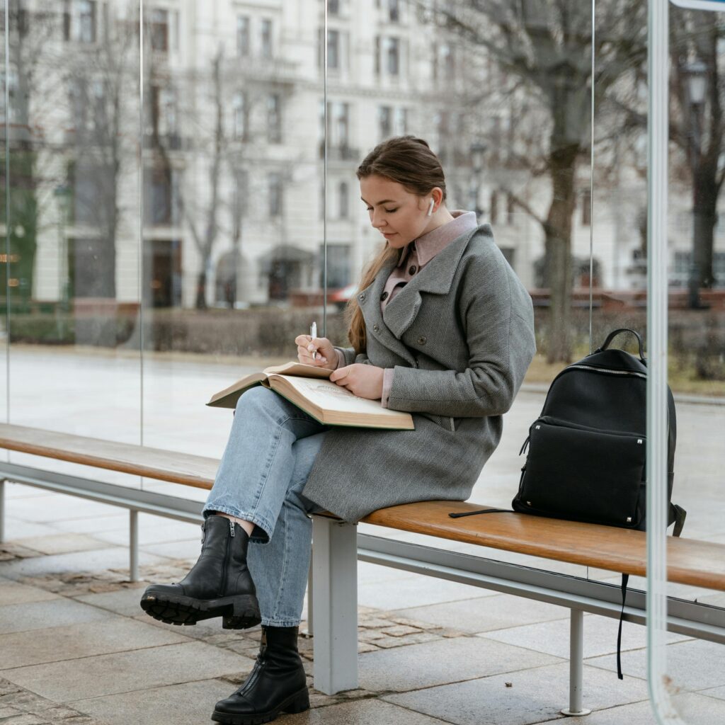 A young student sitting on a bench outdoors during winter, writing in a notebook with a black backpack beside her. The scene captures the essence of a study abroad journey during the January intake, blending focus and adaptability to new environments.